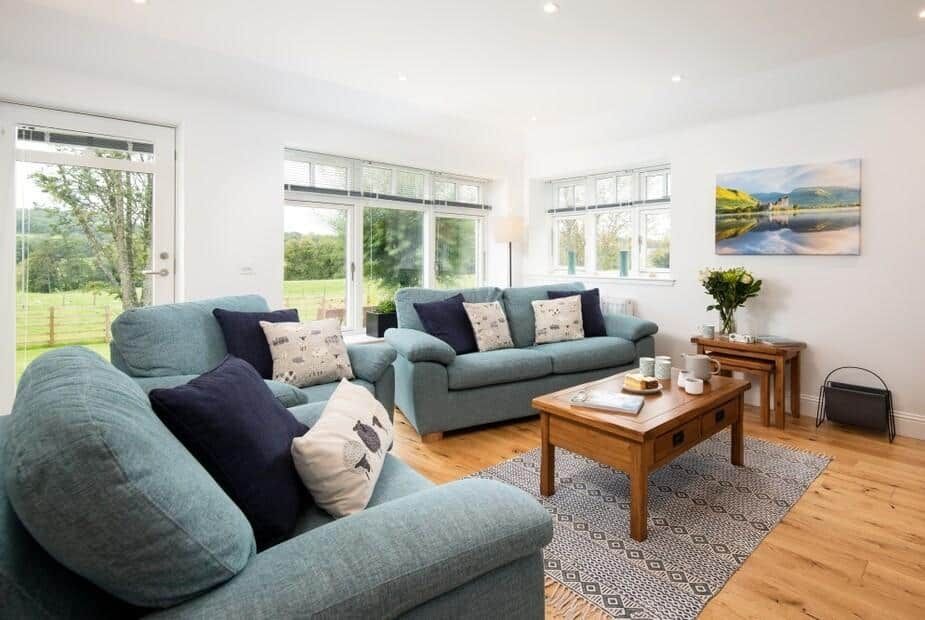 Bright living room with blue sofas and large windows overlooking green fields at Gaidrew Cottage